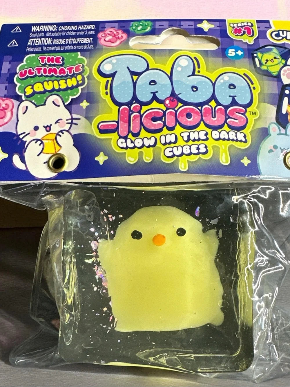 A packaged Taba-Licious Glow in the Dark Cube toy featuring a small yellow chick-shaped squishy inside a dark cube, sealed in clear plastic under a colorful branded card.