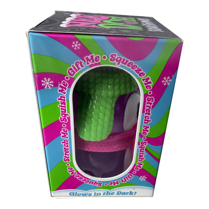 Sex toy packaging box featuring a green textured, snake-like dildo with a purple base and pink strap, visible through a clear window. The box has a colorful pink, green, and blue retro design with text reading 'Gift Me', 'Squeeze Me', 'Stretch Me', and 'Glows in the Dark!'.
