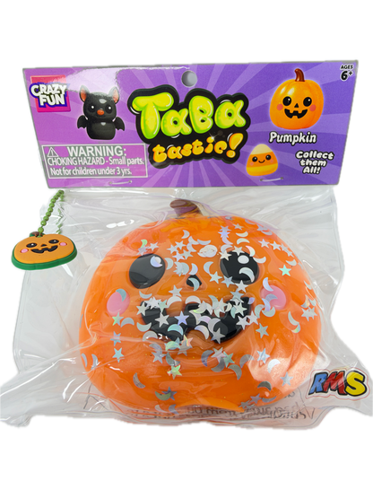 An orange squishy pumpkin toy in clear plastic packaging next to its purple retail card, featuring a carved pumpkin face, black cat eyes, and silver star glitter.