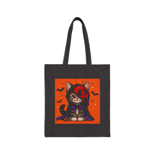 A black canvas tote bag with long handles, featuring a square graphic print of a cute cartoon cat dressed in a red Ghostface mask and purple cape against an orange background with bats and stars.
