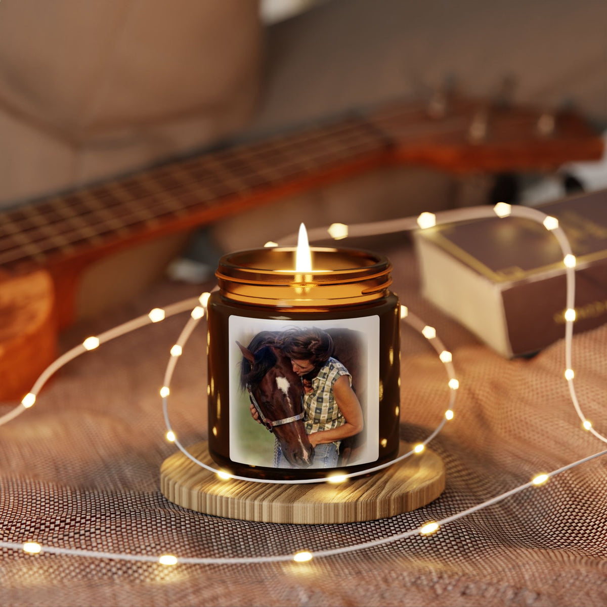 A lit amber glass jar candle sits on a small wooden coaster on a textured blanket, surrounded by warm white fairy lights. The candle features a custom printed photo of a woman hugging a horse on the front label.