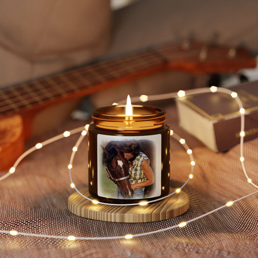 A lit amber glass jar candle sits on a small wooden coaster on a textured blanket, surrounded by warm white fairy lights. The candle features a custom printed photo of a woman hugging a horse on the front label.