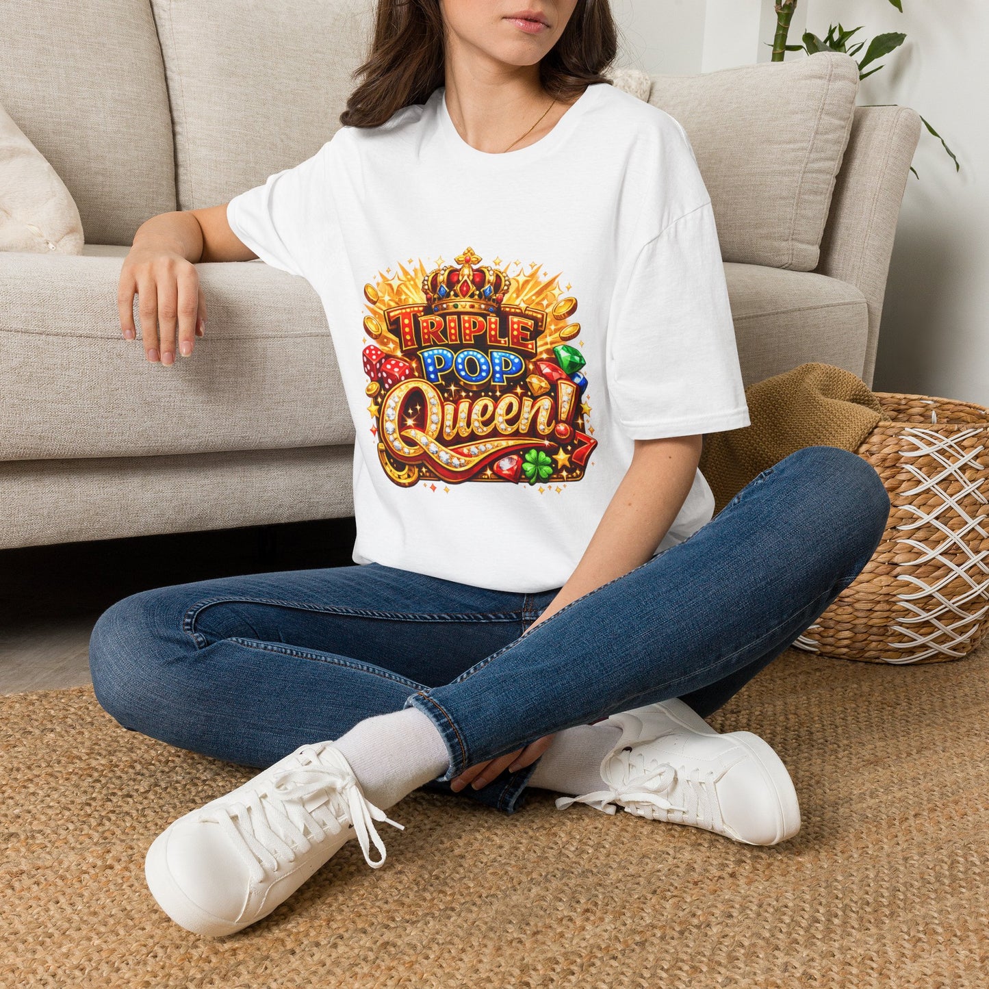 A woman sitting cross-legged on a woven rug in front of a beige sofa, wearing a white graphic T-shirt with a colorful 'Triple Pop Queen' design, blue jeans, and white sneakers.