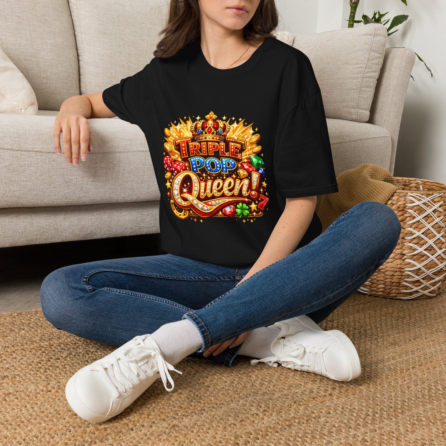 A woman sitting cross-legged on a woven rug in front of a beige sofa, wearing a black graphic T-shirt with a colorful 'Triple Pop Queen' design, blue jeans, and white sneakers.