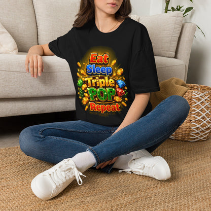 A woman sitting cross-legged on a woven rug in front of a beige sofa, wearing a black graphic T-shirt with colorful text reading 'Eat Sleep Triple POP Repeat' and a bright gem-and-gold design.