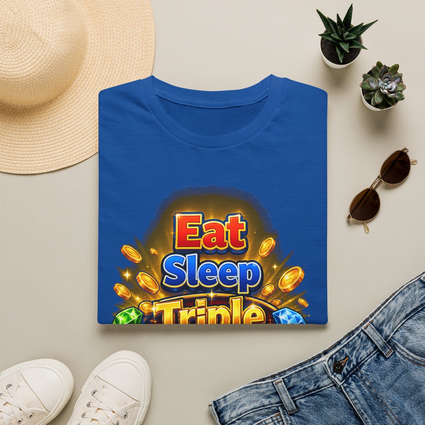 A folded blue T-shirt with a colorful 'Eat Sleep Triple' graphic print, styled in a flat lay with a straw hat, white sneakers, denim shorts, sunglasses, and small potted succulents on a light beige background.