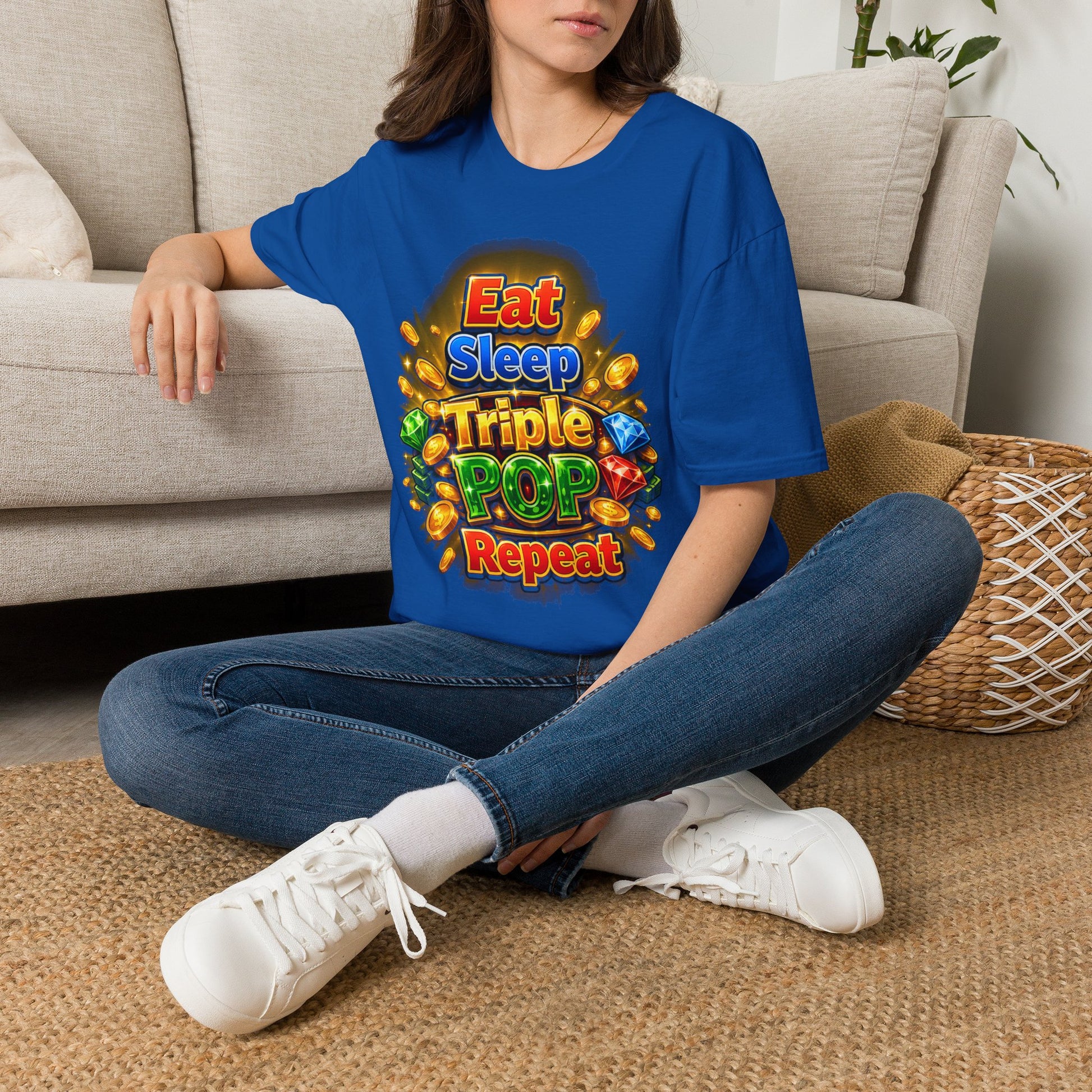 A woman sitting cross-legged on a woven rug in front of a beige sofa, wearing an oversized royal blue t-shirt with a colorful 'Eat Sleep Triple POP Repeat' graphic print, paired with blue jeans and white sneakers.