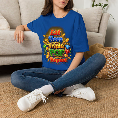 A woman sitting cross-legged on a woven rug in front of a beige sofa, wearing an oversized royal blue t-shirt with a colorful 'Eat Sleep Triple POP Repeat' graphic print, paired with blue jeans and white sneakers.