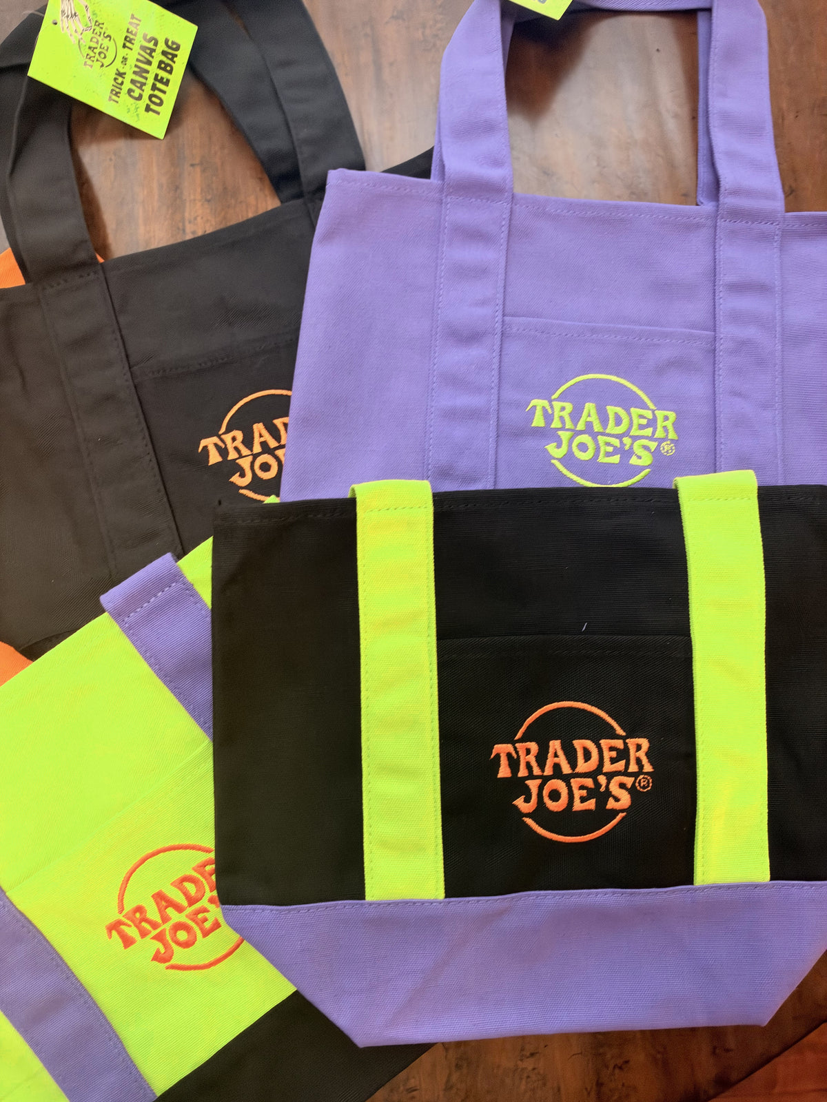 A purple and black canvas tote bag with bright neon yellow-green side stripes and a prominent orange Trader Joe's logo on the front pocket, displayed on a wooden surface with other similar bags partially visible.