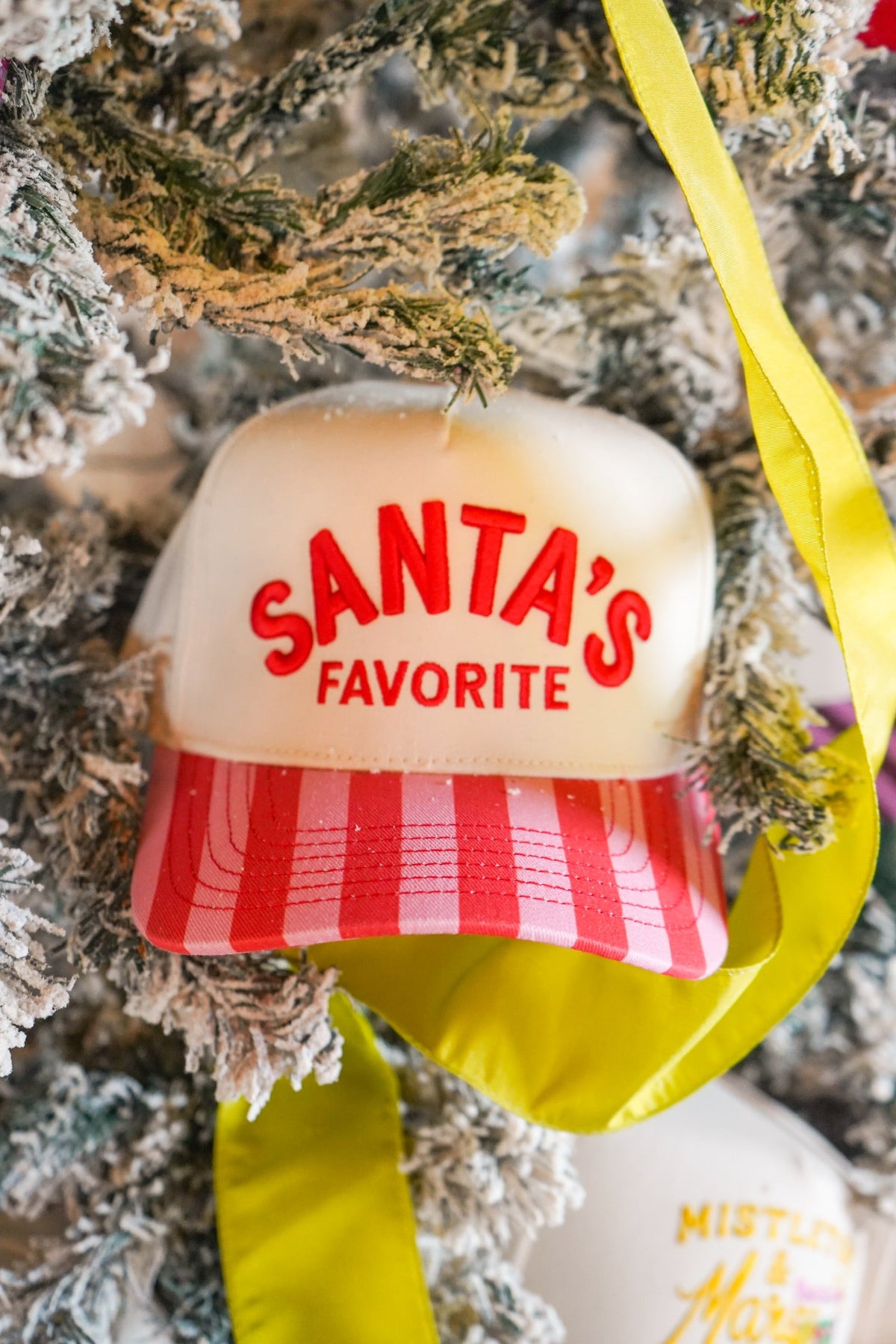 A Christmas trucker hat . The hat is cream-colored with red text reading 'SANTA'S FAVORITE' and a red-and-white striped brim.