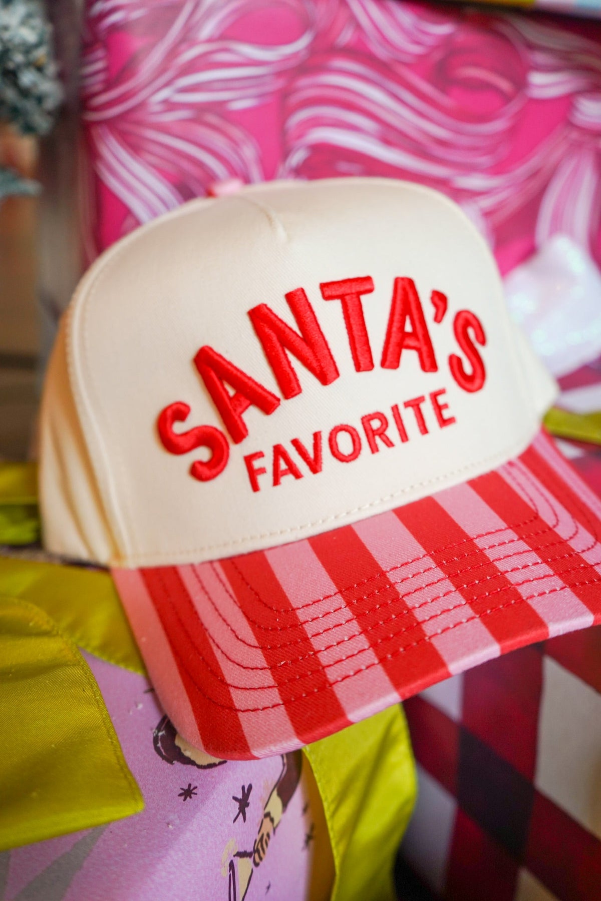 A close-up of a Santa's Favorite trucker hat with a cream crown, red embroidered text, and a red and white striped hat, set against a colorful, festive background.
