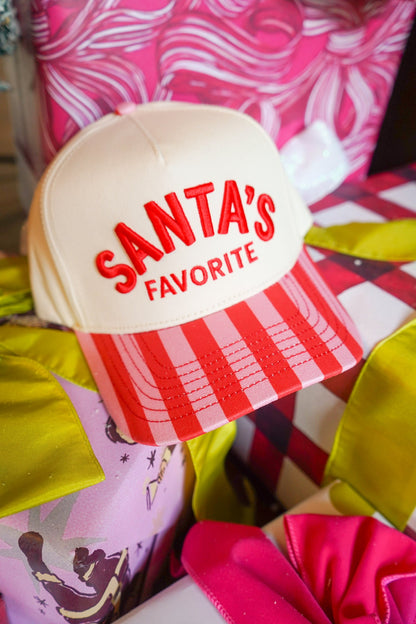 A Santa's Favorite trucker hat with a cream crown, red embroidered text, and a red-and-white striped visor, nestled among colorful gift boxes.