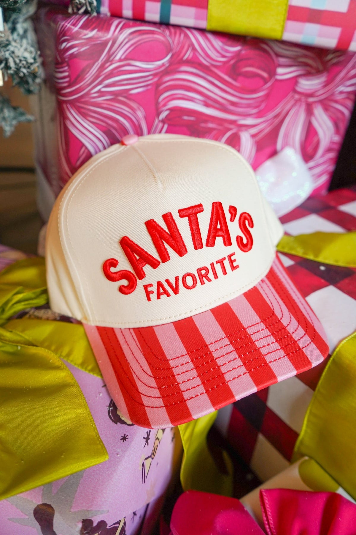 A cream-colored baseball cap with red embroidered text reading 'SANTA'S FAVORITE' and a red-and-white striped brim, styled on top of colorful wrapped gift boxes.