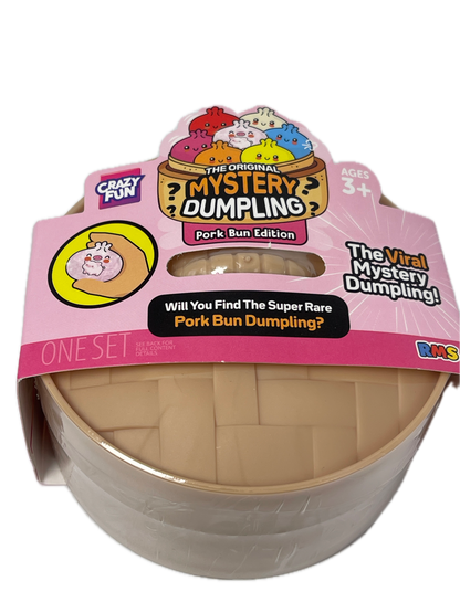 A toy product in a clear plastic dome package with a pink retail card. The item inside appears to be a beige textured cylindrical toy or container labeled 'The Mystery Dumpling' and 'Pork Bun Edition.