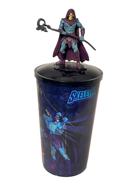 A Skeletor action figure emerging from a printed display cup featuring blue and purple comic-style artwork and the word 'SKELETOR' on a white background.