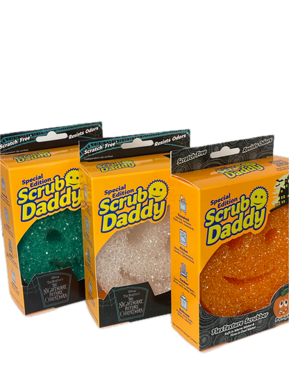 Three packaged Halloween-themed Scrub Daddy products stacked vertically on a white background. The packages are orange with black and yellow branding, showing the scrubby inside: a textured orange scrubby at the top, a translucent white/clear scrubby in the middle, and a dark green scrubby at the bottom.