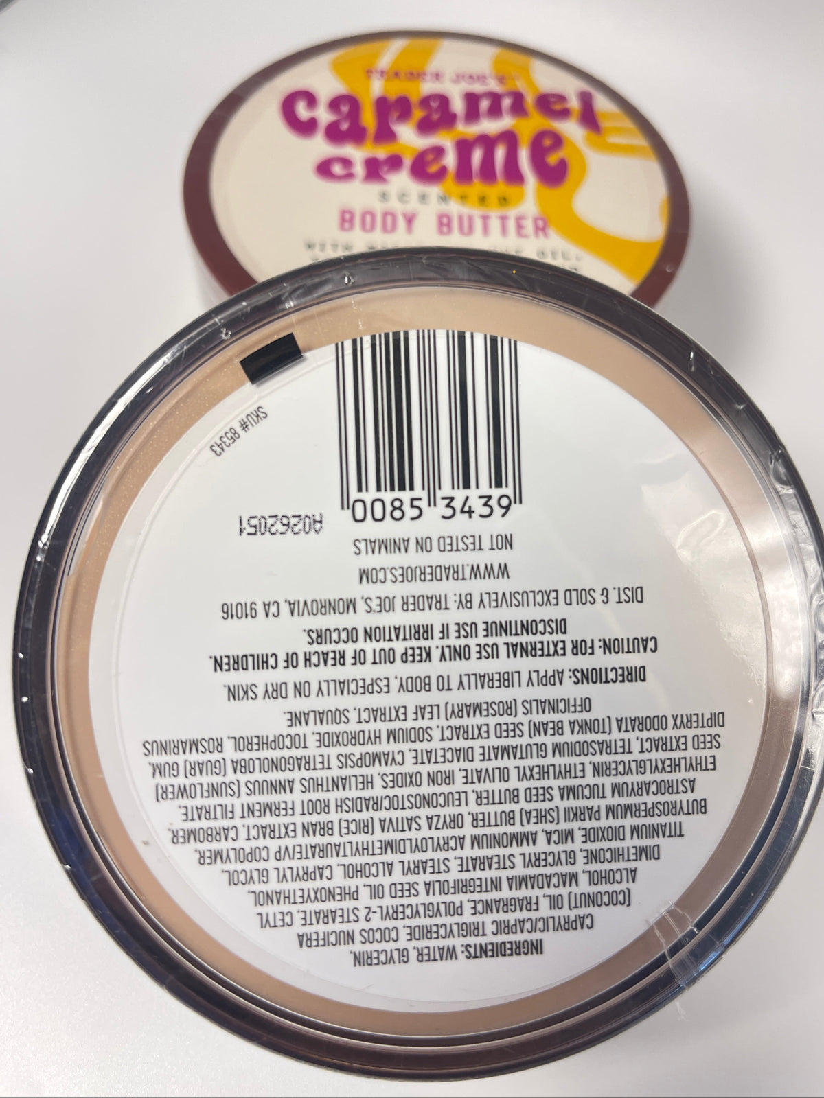 A close-up of the bottom of a round jar of body butter, showing the printed barcode label and product text. A second jar labeled 'Caramel Creme Body Butter' is partially visible in the background.