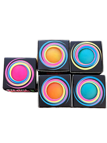 Four square black cosmetic boxes arranged on a dark wooden surface, each featuring a colorful concentric ring design in pink, yellow, blue, and green around a bright circular center. One box is open, showing a pink product inside.