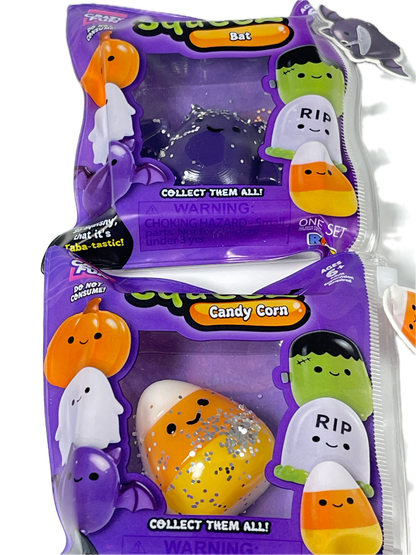 Two packaged collectible squishy toys in clear plastic bags with purple Halloween-themed packaging. One bag contains a dark purple ghost squishy, and the other contains a candy corn squishy with orange, white, and silver glitter details.
