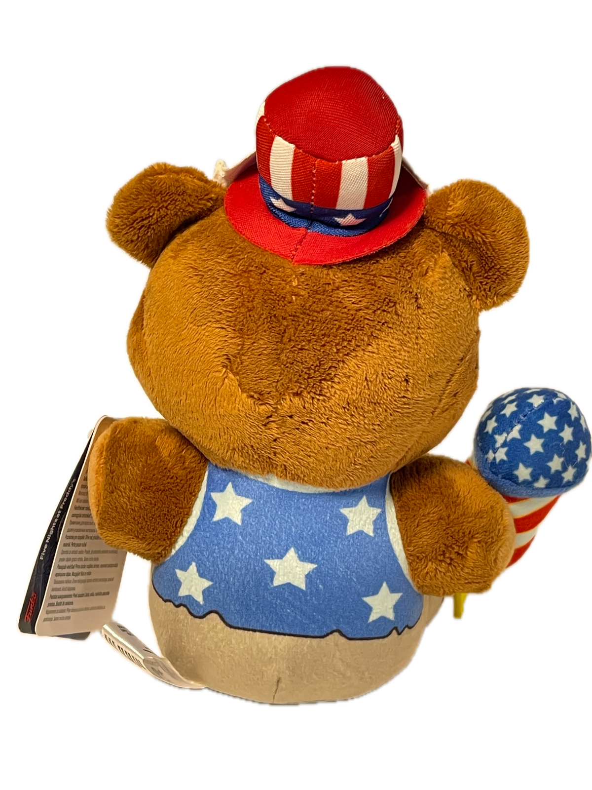 A brown plush teddy bear wearing a patriotic red, white, and blue outfit with stars and stripes, shown on a plain white background with a hangtag attached.