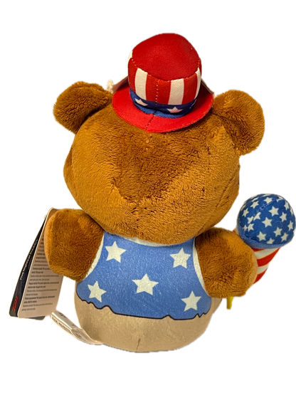 A brown plush teddy bear wearing a patriotic red, white, and blue outfit with stars and stripes, shown on a plain white background with a hangtag attached.