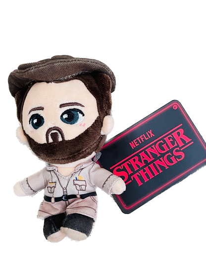Plush toy of a character from 'Stranger Things' holding a sign with the show's logo on a white background.