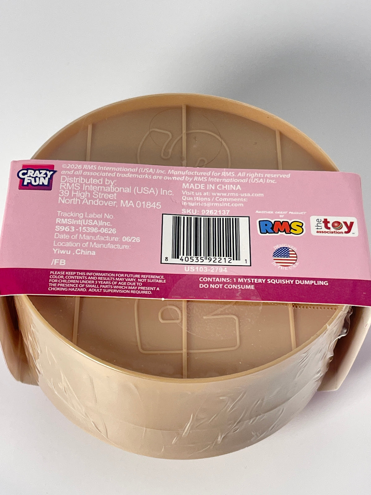 A beige plastic toy bowl wrapped in clear plastic and covered with a pink product information label, photographed on a light gray background.