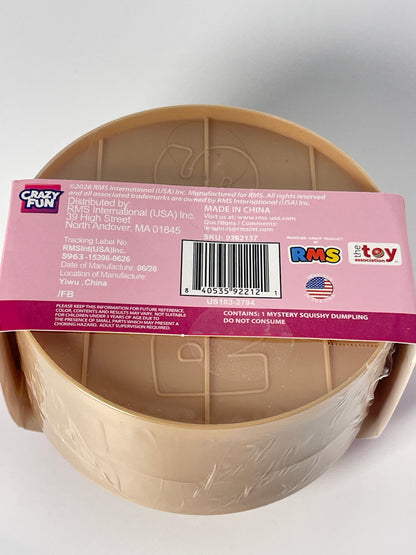 A beige plastic toy bowl wrapped in clear plastic and covered with a pink product information label, photographed on a light gray background.