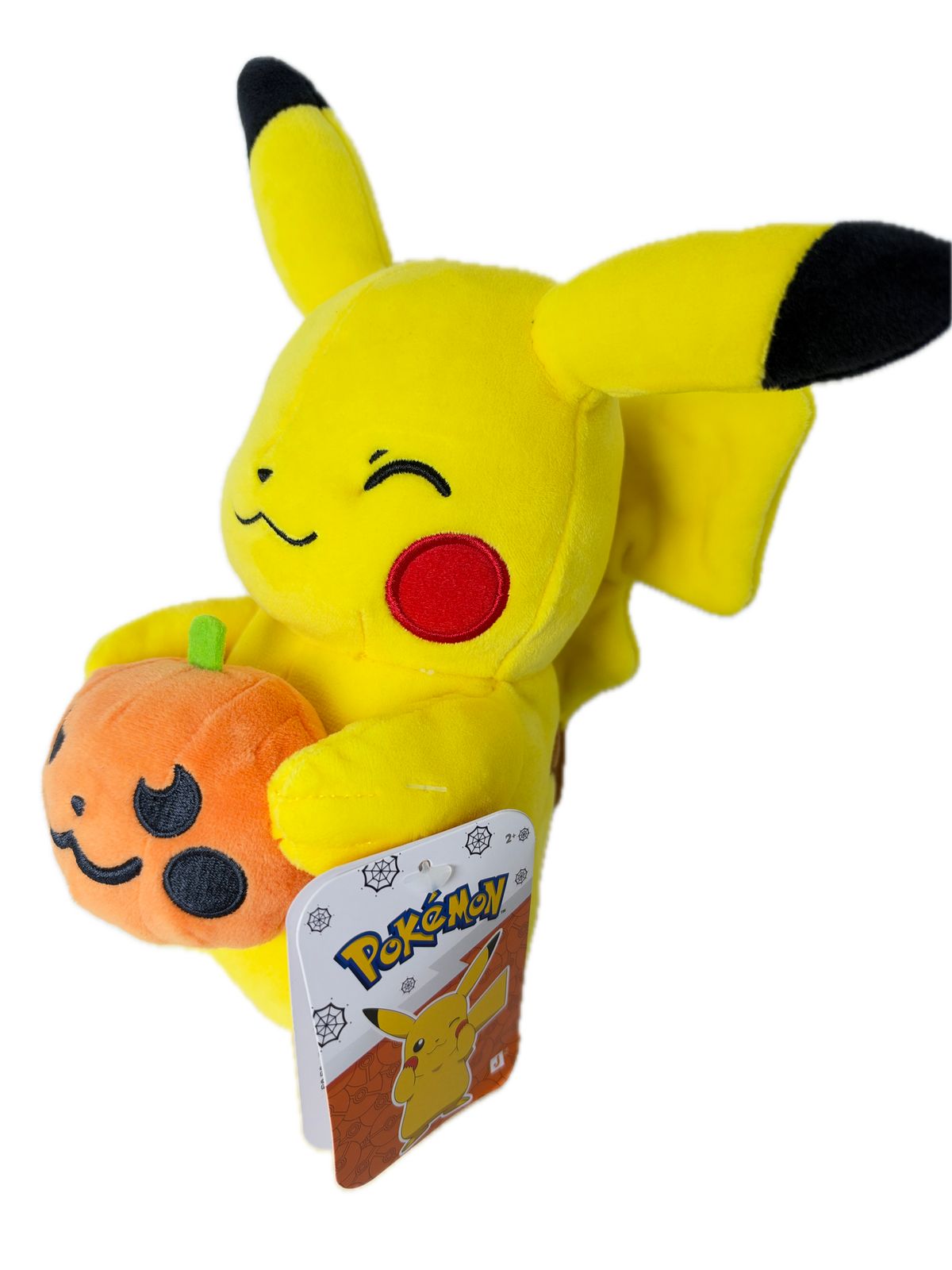 A yellow Pikachu plush toy lying on its side, holding an orange pumpkin with black facial features. The plush has black-tipped ears and tail tips, a red circular cheek emblem, and a visible Pokémon tag attached near its head.