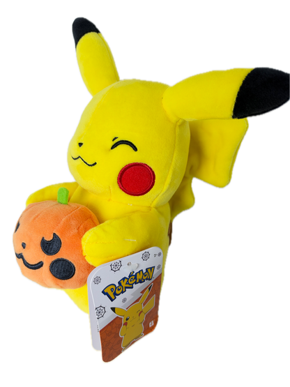 A yellow Pikachu plush toy lying on its side, holding an orange pumpkin with black facial features. The plush has black-tipped ears and tail tips, a red circular cheek emblem, and a visible Pokémon tag attached near its head.