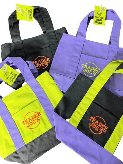 Three Trader Joe's canvas Halloween tote bags in purple, black, and neon yellow-green, arranged on a wooden surface. Each bag features the orange Trader Joe's logo on the front pocket and has a yellow hangtag attached.