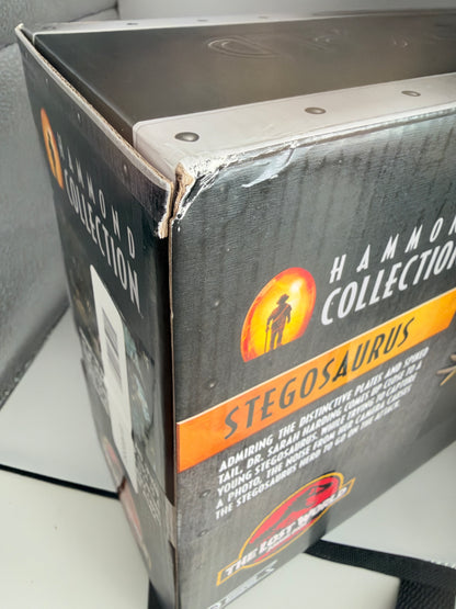 A close-up of a boxed collectible item labeled 'Hammerhead Collection Stegosaurus' with a black box, orange and yellow graphics, and visible wear at the top edge.