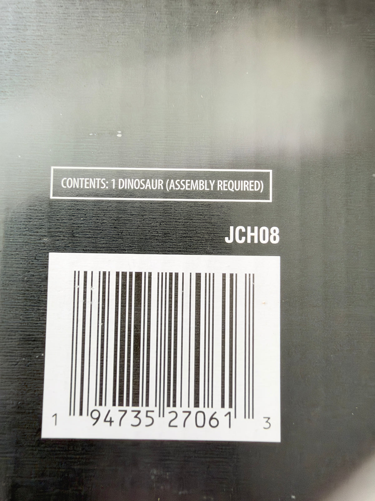 Close-up of the back of a black product box showing a barcode, the text 'CONTENTS: 1 DINOSAUR (ASSEMBLY REQUIRED)', and the code 'JCH08'.