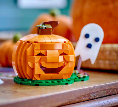 A LEGO-style orange jack-o'-lantern set on a green base, photographed on a wooden surface with blurred pumpkins and a white ghost decoration in the background.