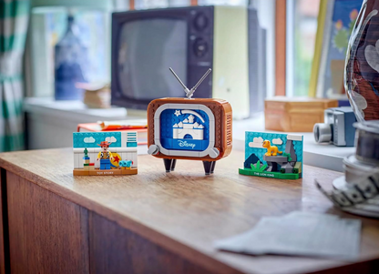 A retro-style television toy set with two small Disney-themed building block scenes displayed on a wooden table in a softly blurred room setting.