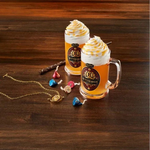 Two glass mugs with whipped cream and a logo on a wooden surface, surrounded by jewelry and candies.