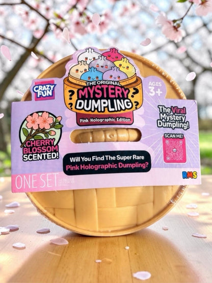 A packaged toy product labeled 'The Original Mystery Dumpling' in pink holographic edition, shown in a basket-like container with a cherry blossom themed background and scattered petals.