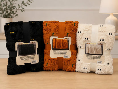 Three folded plush throw blankets in black, orange, and cream are displayed side by side on a light wood surface. The black blanket has yellow cat eyes, the orange blanket has small green pumpkin details, and the cream blanket has ghost faces.