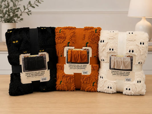 Three folded plush throw blankets in black, orange, and cream are displayed side by side on a light wood surface. The black blanket has yellow cat eyes, the orange blanket has small green pumpkin details, and the cream blanket has ghost faces.